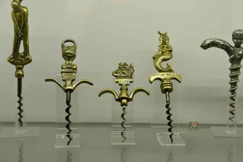 一排五個造型精美的復古開瓶器，置於透明壓克力底座上，背景為純白色。開瓶器的把手由黃銅和銀製成，造型各異，包括：  一位揮桿的高爾夫球手。  一個風格化的貓頭或石像鬼頭。  一艘標示著「A. Galleon 1588」的帆船。  一條標有“Torquay Mermaid”字樣的美人魚。  一個銀色螺旋形把手，由一個小天使造型構成。