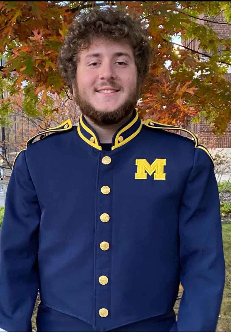 一名身穿藍色軍樂隊制服的男子，左胸印有亮黃色字母「m」。