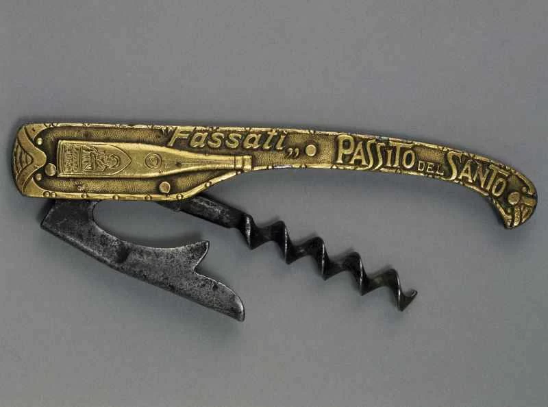 圖中展示的是復古折疊侍者開瓶器（卡爾維恩克設計），背景為中性灰色。手柄採用壓花金色金屬製成，飾有「Fassati Passito del Santo」字樣和酒瓶圖案。手柄展開後，可見深色金屬螺旋鑽頭和帶有凹槽的銀色槓桿臂，展現了早期便攜式開瓶器的機械設計。
